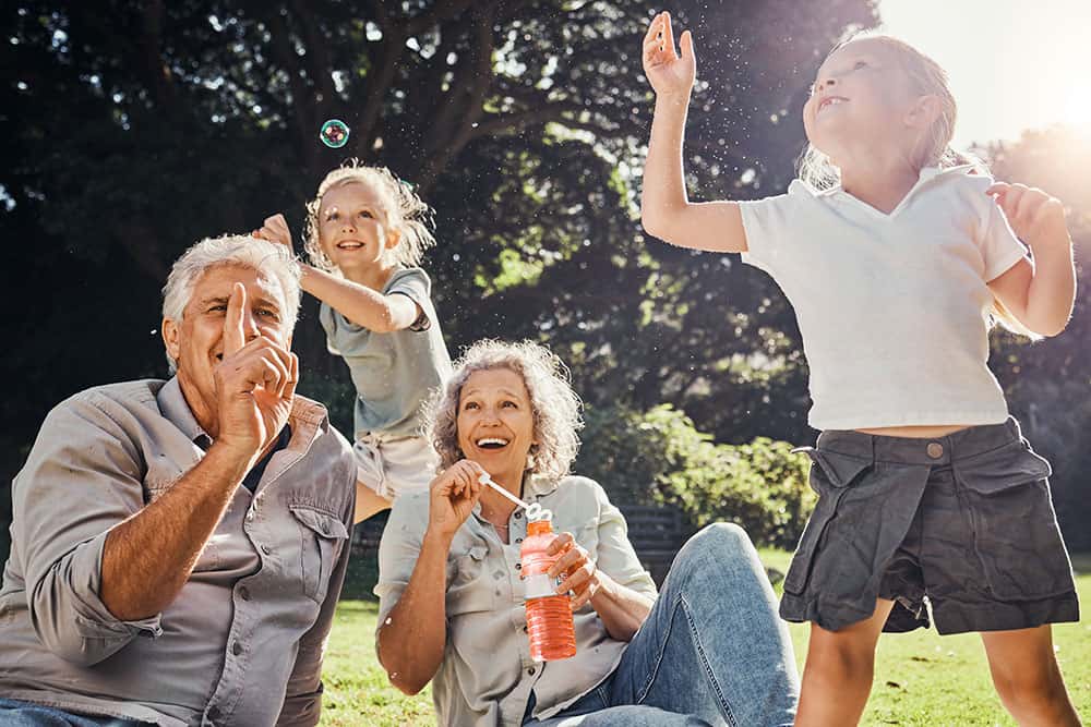 Grandparents sitting in the grass while two granddaughters blow bubbles